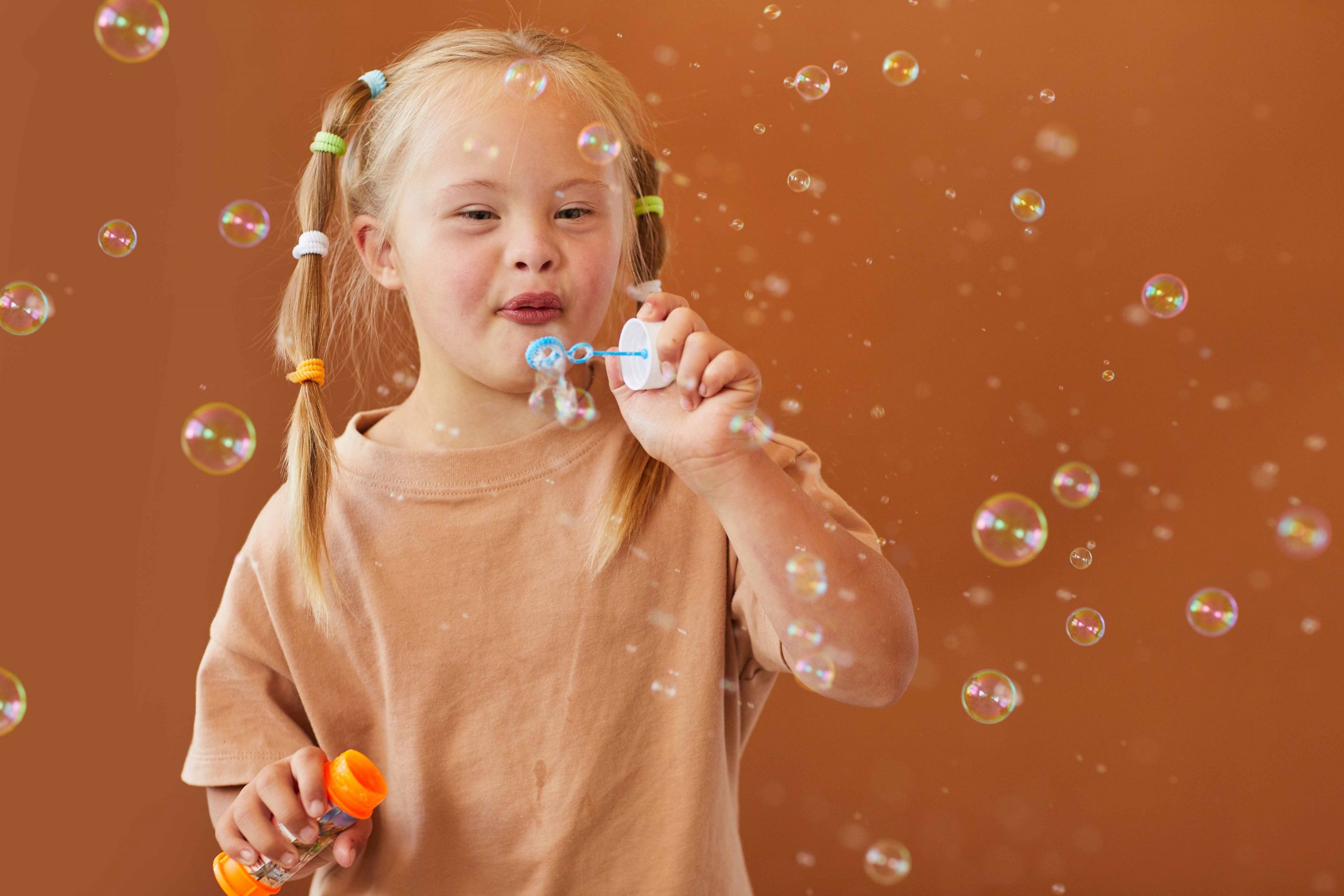 Waist up portrait of cute girl with down syndrome blowing bubbles while posing against brown background in studio, copy space