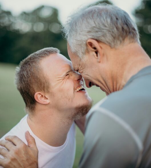 A smiling young man and an older man touch foreheads and laugh together outdoors, showing affection and joy in a grassy park setting.