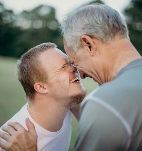 A smiling young man and an older man touch foreheads and laugh together outdoors, showing affection and joy in a grassy park setting.