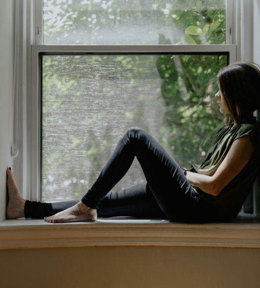 A person sits quietly on a windowsill, gazing out at the greenery outside as natural light fills the room.