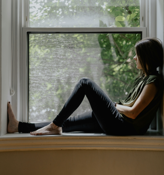 A person sits quietly on a windowsill, gazing out at the greenery outside as natural light fills the room.